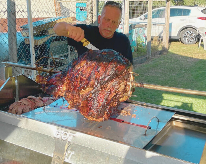Man grilling a large roasted meat on a metal grill outdoors.